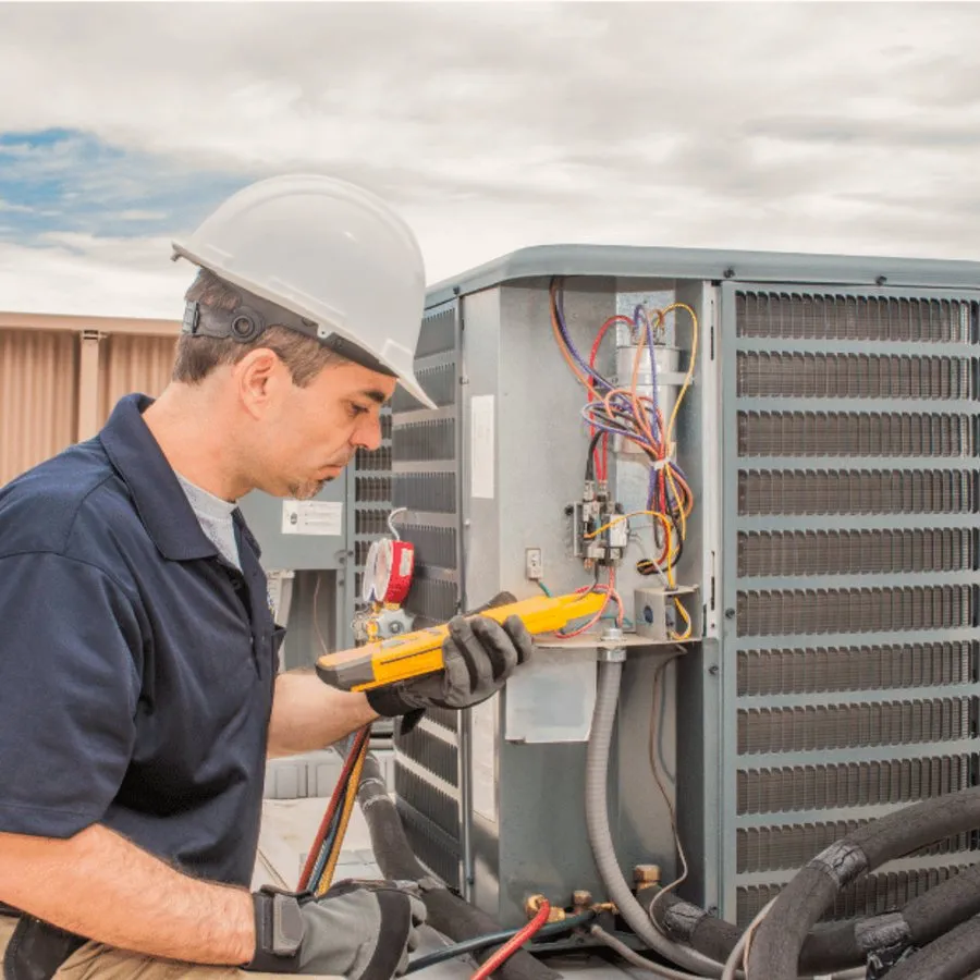 HVAC technician in a hard hat testing an outdoor air conditioner with a multimeter